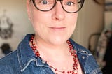 Woman with chemo hair growing out, wearing glasses, a red necklace and a denim jacket