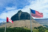 Photo of a Canadian and American flag flying side by side in front of a mountain