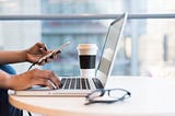 A laptop, a styrofoam cup and glasses on what seems to be a round wooden table. There are black feminine manicured hands; the left holding a charging phone and the right one on the laptop.