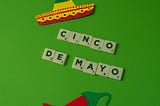 Cinco de Mayo celebration decorations on a bright green background featuring letter tiles spelling ‘CINCO DE MAYO’, a colorful sombrero cutout, and a red chili pepper cutout.