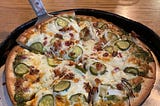A pan of pizza with chopped dill pickles, bacon and onions.