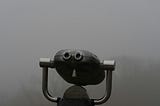 A pair of gray, metal, coin-operated tower viewers on a stand. The background behind them is completely obscured by a thick, dense fog, making it impossible to see into the distance.