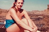 A brown-haired teenage girl wearing a blue full-piece bathing suit sits on a rocky landscape, her arms hugging her legs to her chest. She looks down with an unsure look on her face. The beach is far off in the distance.