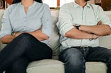 A man and woman are seated and can only be seen from the waist down. They are both wearing a light gray shirt and blue jeans.