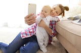 Mother holding a baby and taking a photo of them both.