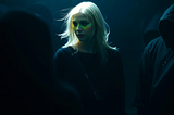 The image shows a digitally created woman with pale blonde hair standing in a dark, shadowy environment. She is illuminated by a soft light, making her stand out as a unique entity among the crowd. The figures around her are wearing dark hooded clothing, blending into the shadows, which enhances the sense that she is distinct and special. This scene symbolizes the idea that every one of us is unique and special in the vast universe.