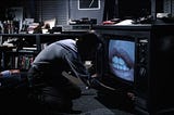 An image from David Cronenberg’s Movie “Videodrome,” featuring the main character crouched in front of a television, apparently possessed by his love interest.