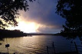 Bright and dark clouds over calm waters with overhanging cedar branches in the foreground.