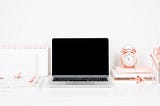 A Macbook, notebooks and a rose gold clock on a white desk