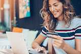 Young woman holding a credit card while sitting in front a laptop with a smile.