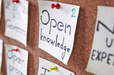 Yellow post-it notes attached to a cork board with colorful push-pins. The fully visible notes say (from ascending to descending), “Education ? ? ?,” “Open Knowledge,” and “Team” crossed out with “Community” written below.