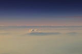 A deep blue sky above wispy white clouds, as if seen from an aeroplane. Mountain tops can just be seen through the clouds in the distance.