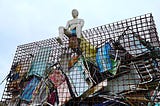 concrete gray sculpture of a man in swimming trunks perches high atop a giant metal cage filled with discarded beach chairs, the cage is 30 feet (10m) high so he is silhouetted against the sky