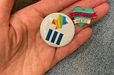 A pin with the shape of Texas filled in with rainbow colors and another pin with she/her/hers pronouns.