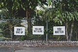 Chain link fence with three white signs with black letters saying Don’t Give Up, You Are Not Alone, You Matter.