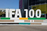 The image shows a large, three-dimensional text installation that reads “IFA 100” in white letters. It is placed outdoors, likely in front of a building with banners mentioning “IFA” and other partially visible text. The numbers “100” suggest a centennial celebration or anniversary. The setting appears to be a sunny day with clear skies.