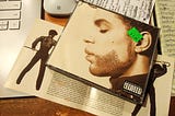 My copy of Prince’s The Hits/The B-Sides (with its $9.99 price tag visible) sits on top of its folded out booklet. A couple images of Prince standing up and holding a cane can be seen from beneath the CD package on top. My handwriting-scribbled notebooks, laptop, and mouse peek into the frame towards the edges.