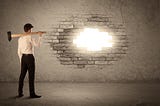 a man with a sledge hammer stands in front of a brick wall. He has knocked a small hole in the wall and sunlight is shining through.