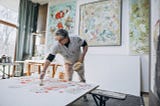 A man with greying hair paints a canvas laying flat in front of him with abstract paintings on the wall behind him.