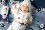 An infant wears a white spacesuit-looking sleeper and lays against a surface-of-the-moon themed blanket. A toy rocket lies on its side next to the baby.