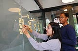 A woman in a light blue sweater places a sticky note on an idea board. A man with glasses stands beside her.