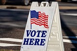 A picture of a small polling place sign instructing voters to “Vote Here.”