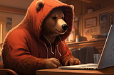 A brown bear wearing an orange hoodie sitting at a desk while looking at a MacBook