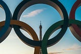 An image of the Olympics symbol on a clear day in paris with the Eifel Tower displayed in the middle.