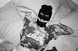 A grayscale photo of a woman lying on a bed while wearing a 3-hole full-face ski mask