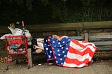 A houseless man sleeps on a bench next to a shopping cart full of his belongings, draped in an American flag blanket.