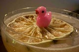 A light yellow cocktail in a clear glass topped with a dried citrus wheel, with a small pink plastic duck figurine sitting on the citrus slice.