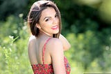 An attractive girl with dark hair is looking back over her shoulder to the camera. The shot is from her waist up, showing her wearing a summer dress.