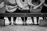 Black and white photographs of five people sitting on a bench facing away from the viewer. The people have their arms around each others back.