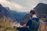 A digital nomad sitting on the grass, on top of a mountain working on his laptop