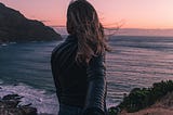 Two people holding hands. The woman facing towards the sea, looking out to the horizon.