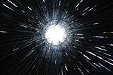 A photo from inside an art installation made of long black stems of bamboo arranged in a circle. The photo is taken from the ground looking up through the circle opening at the top and into a bright, white, overcast sky. Several small gaps between the stems are creating the effect of small lines of white light pointing into the centre white circle.