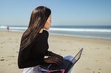 Woman in black long sleeve shirt using black laptop pink phone case working on computer at the beach looking out in the distance remote work