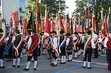 Austrian men wearding lederhosen standing on the road and holding different type of flags that represent thier villages or town.