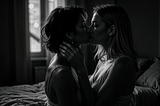 Two Caucasian women, one with wavy chestnut short hair and the other with sleek straight blonde hair, deep kissing in a dimly lit bedroom with shadows dancing on the walls. One of the women is gently touching the other’s face.