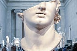 A marble bust of a partially damaged ancient statue displayed in a museum, showing the lower part of the face and neck. The background features a museum hall with blurred visitors and other sculptures.