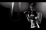 A woman wearing boxing gloves, a sports bra, and leggings, is ready to punch a heavy bag.