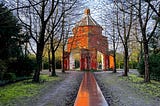 A soggy winter’s day in a public park, bare trees line a slick walkway that leads to a red brick rotunda, the lower level has high opan arches and tiny figures are walking through
