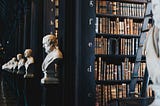An old library filled with large books and white marble busts of historic men