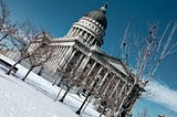 angled photo of Utah State Capitol building — Neoclassical architecture with Corinthian columns around the outside and one level below the black dome. It is winter with snow on the ground and several trees without leaves.