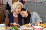 A young Chinese American woman rests her head against the shoulder of an elderly Chinese woman