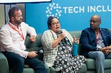 Three people sit on a couch in front of a blue Tech Inclusion sign, discussing inclusion ecosystem building for a panel.