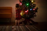 Little girl in a checkered dress standing beside the Christmas tree.