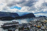 A large cruise ship docked next to the Norwegian town of Alesund. To the right of the image are hundreds of multi-colored buildings. The cruise ship is in a semi-circular bay and mountains are visible in the background.