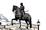 Charles I statue standing at the head of Whitehall, just below Trafalgar Square — image shows a bronze man on a bronze horse on a stone plinth, with the tops of buildings visible behind, plus the top of a streetlamp