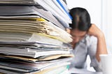 Person sitting at a desk behind a large stack of papers with head in hands.